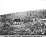 Seven miners, First Nations woman beside log cabin and flume, Hunker Creek