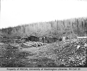 [ Seven miners, First Nations woman beside log cabin and flume, Hunker Creek ]
